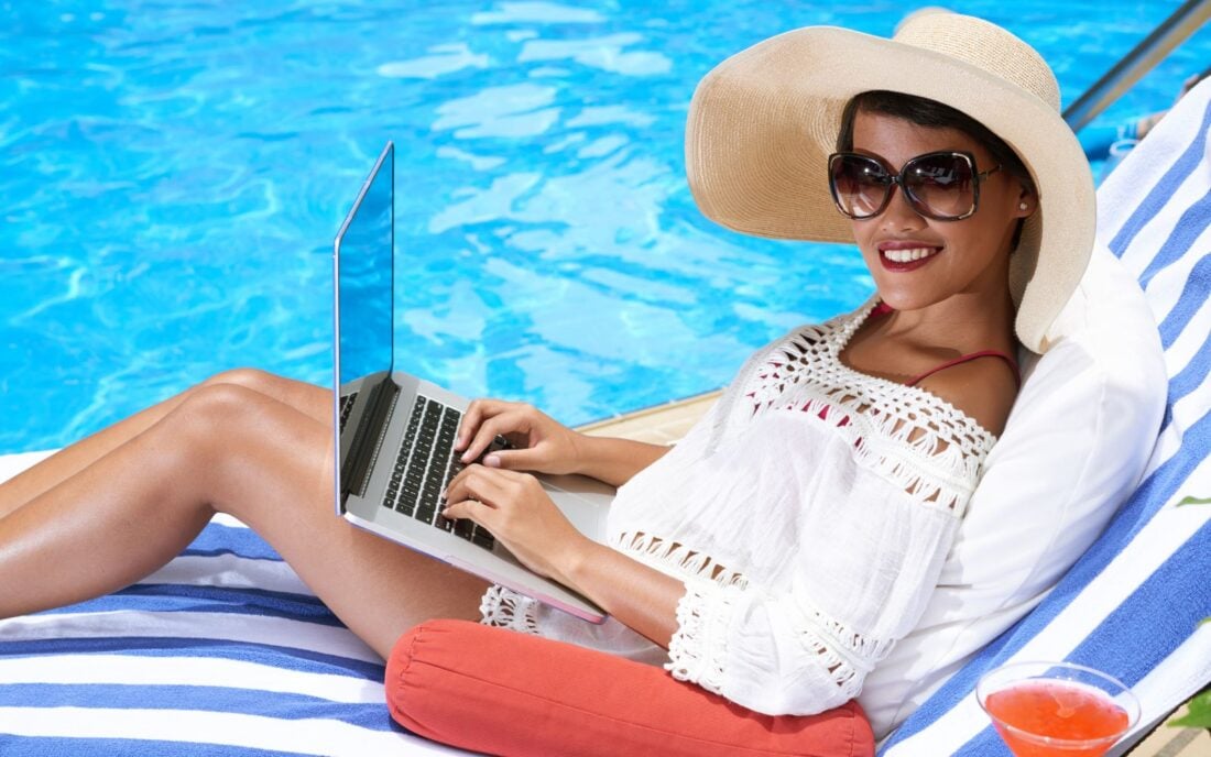 Smiling young woman in straw hat and sunglasses sitting on chaise-lounge and working on laptop as a digital nomad